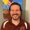 Kevan smiling wearing STAC Spartan head short sleeve shirt with painting of flower and American flag in background