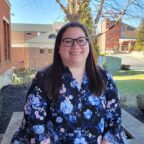 Cassie smiling wearing black rimmed glasses and floral blue shirt with Costello Hall and trees in background