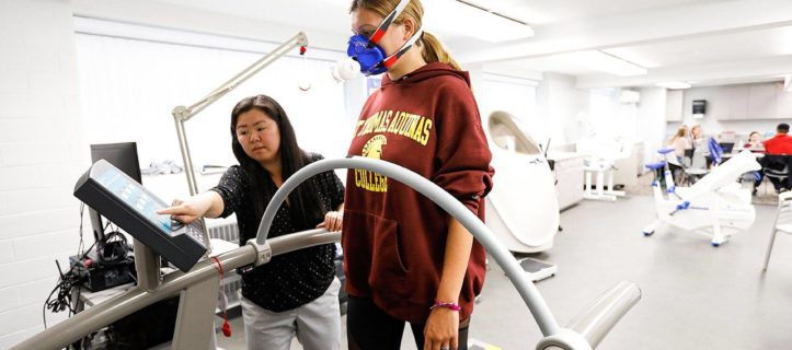 A student and her professor on the treadmill in the exercise lab and students sitting a a desk.