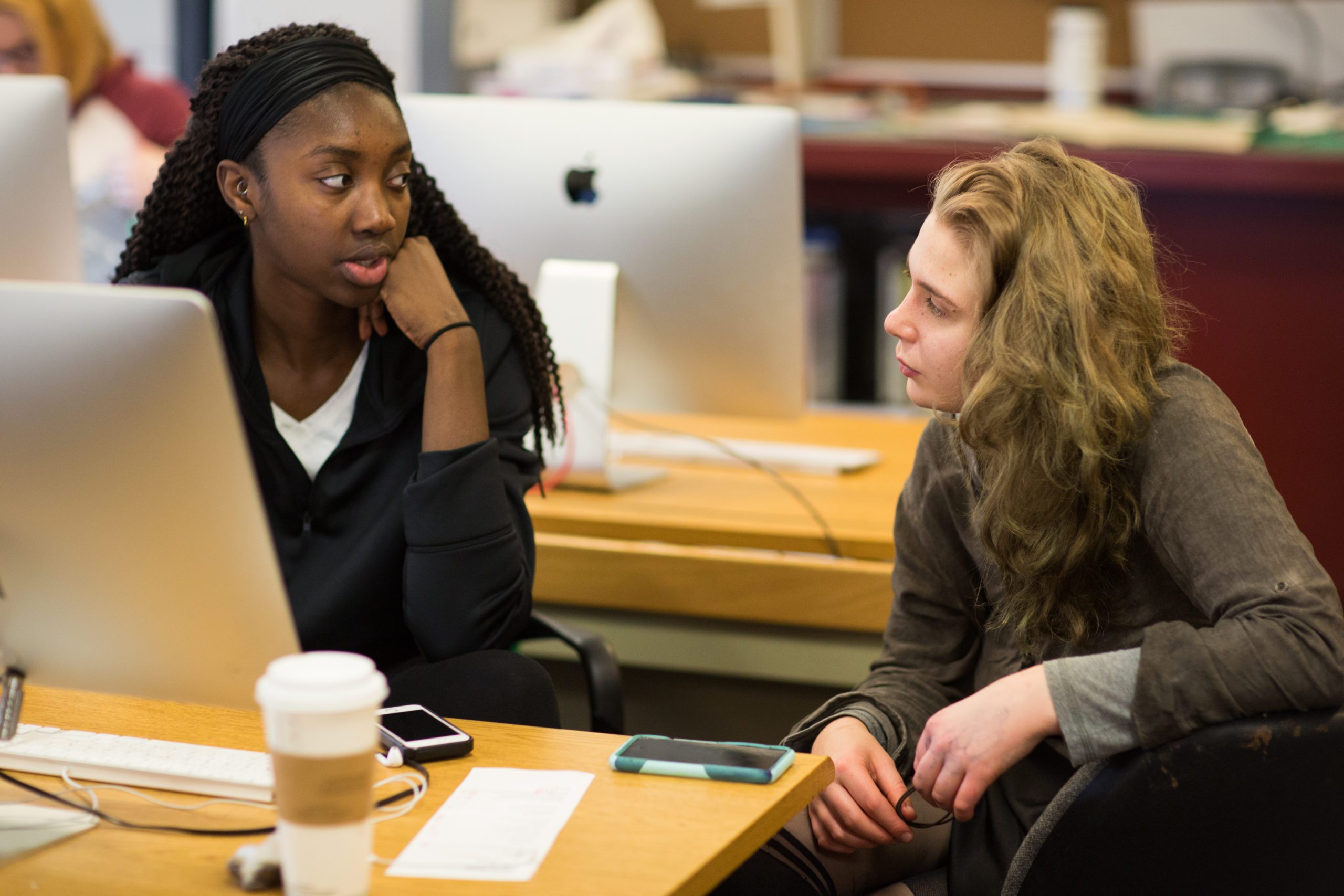 Two female students sitting at a desk in a classroom with computers in front of them.