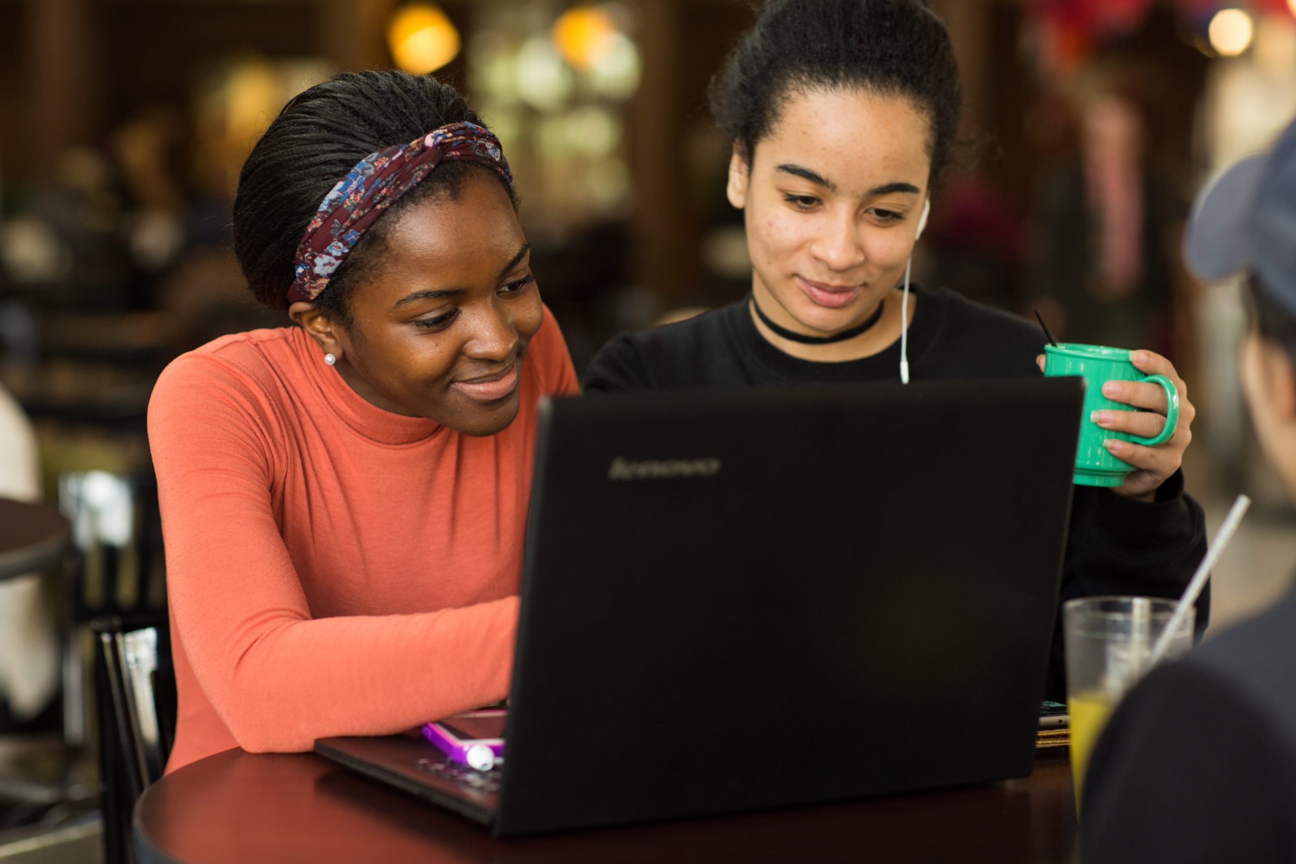 Two female students sitting at a desk with two of them working on a laptop.