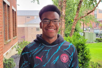 St Christopher smiling wearing glasses and black sweatshirt with borelli hall and green trees and grass in background