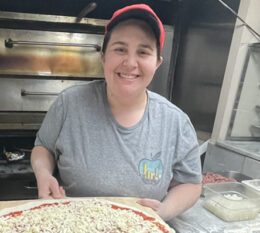 Victoria smiling at work wearing tshirt and hat holding a pizza ready to cook in the oven