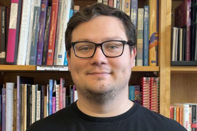 Andrew smiling wearing glasses and black shirt with book stacks in background