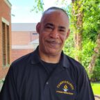 Howie smiling outside wearing Campus Safety black and gold shirt with green tree in background