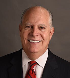 Mark Marotta smiling wearing black suit, white shirt, and red tie