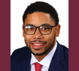 Kameron smiling wearing suit jacket red tie and black rimmed glasses