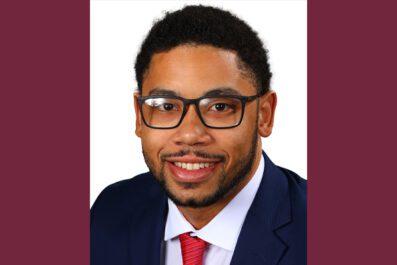 Kameron smiling wearing suit jacket red tie and black rimmed glasses