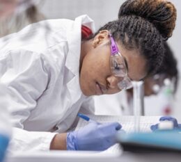 Student in lab with goggles, lab coat, and gloves on looking at data figures on desk
