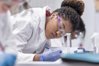 Student in lab with goggles, lab coat, and gloves on looking at data figures on desk